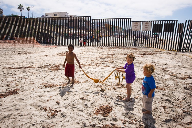 children play a length of seaweed on the beach in front of border wall